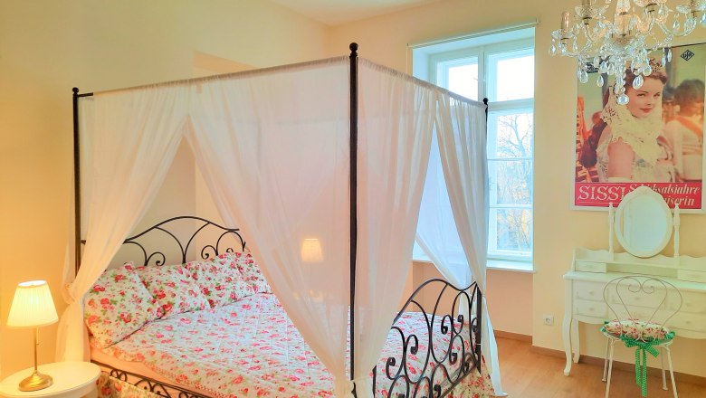 Schlafzimmer mit Himmelbett, © Sisi-Schloss Rudolfsvilla Schlafzimmer mit Himmelbett, Blumenbettwäsche, Kronleuchter und Schminktisch.