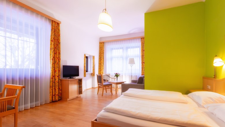 Zimmer mit Hotel Belvedere, © Hotel Belvedere/Gregor Hartmann Helles Hotelzimmer mit Doppelbett, grünem Akzent an der Wand, Fernseher, Sitzbereich und großen Fenstern mit Vorhängen.