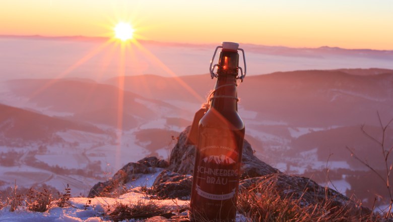 Weissbier aus dem Schneebergland, © Peter Strobl / Karl Tisch Eine Flasche Schneeberg Bräu Bier steht auf einem schneebedeckten Berg bei Sonnenuntergang.