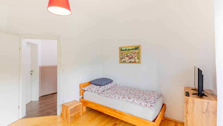 Ferienwohnung Semler, © Wiener Alpen / Christian Kremsl Ein einfach eingerichtetes Schlafzimmer mit Einzelbett, Nachttisch, Kommode und Fernseher. An der Wand hängt ein Blumenbild.