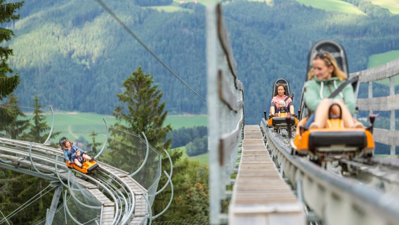 Sommerrodelbahn, © Erlebnisarena St. Corona Menschen fahren auf einer Sommerrodelbahn in den Bergen.