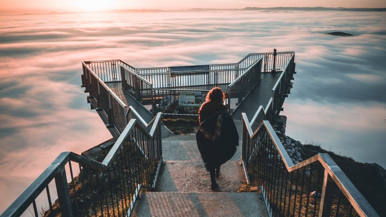 Skywalk Nebel, © (c)NÖW_Pawlikowsky Eine Person geht auf einem Skywalk über einem Nebelmeer in den Sonnenuntergang.