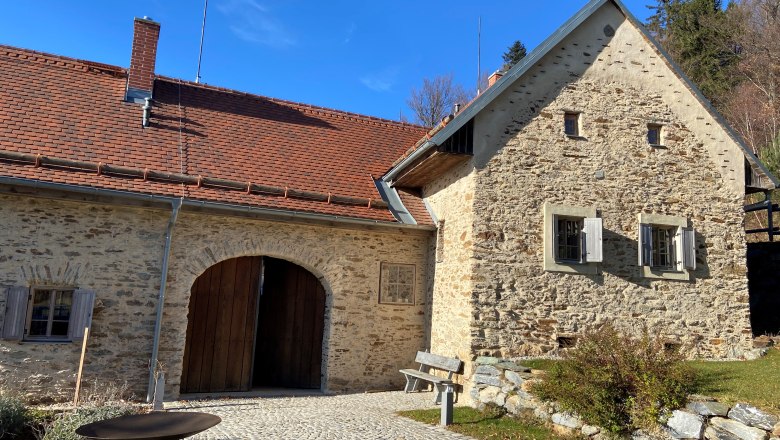 Tailor's goods, © Wiener Alpen Old stone building with red tiled roof and wooden gate, surrounded by paved path and vegetation.