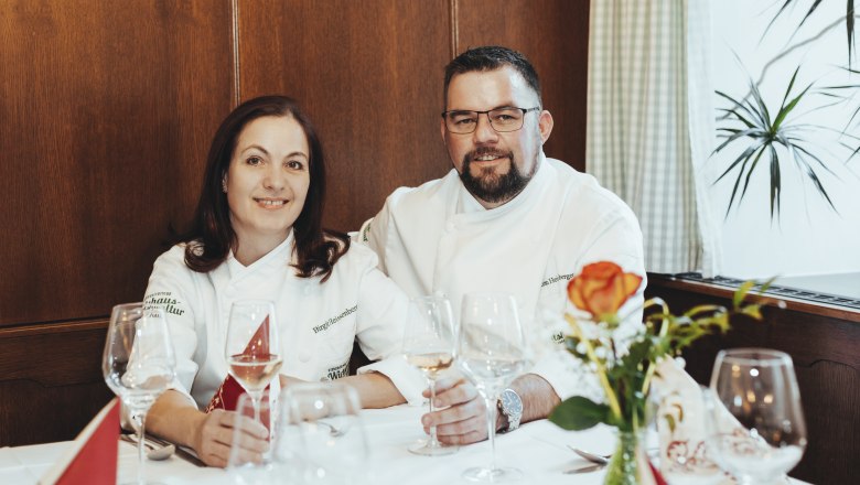 Birgit and Christian Heissenberger, © Niederösterreich Werbung/David Schreiber Two people in chef's jackets sit at a laid table with wine glasses.