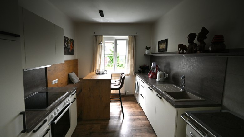 Küche, © Christine Kuppelwieser Moderne Küche mit Holz- und Metallakzenten, Esstisch am Fenster, Küchengeräte und Dekorationen auf Regalen.