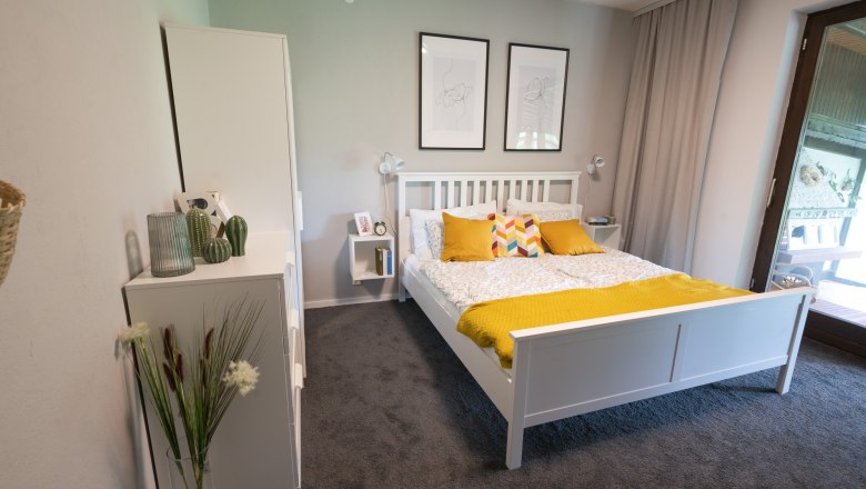 Ferienhaus Schlager Schlafzimmer Erdgeschoss, © Claudia Schlager Schlafzimmer mit weißem Bett, gelben Kissen und Decke, zwei Wandbildern und einem Fenster mit Vorhang.