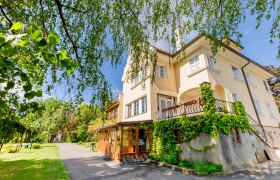 Herrenhaus Woltron, © Wiener Alpen / Christian Kremsl Ein gelbes Herrenhaus mit Efeu bewachsen, umgeben von Bäumen und einem Gartenweg.