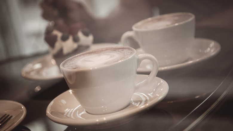 Café specialties, © Hönigmann/Martin Datzinger Two cups of coffee on saucers, a dessert in the background.