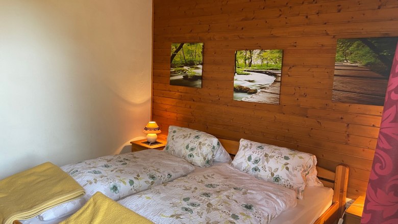 Doppelzimmer, © Wiener Alpen Gemütliches Doppelzimmer mit Holzwänden, einem Doppelbett mit Blumenbettwäsche und gelben Decken, sowie einer Nachttischlampe.