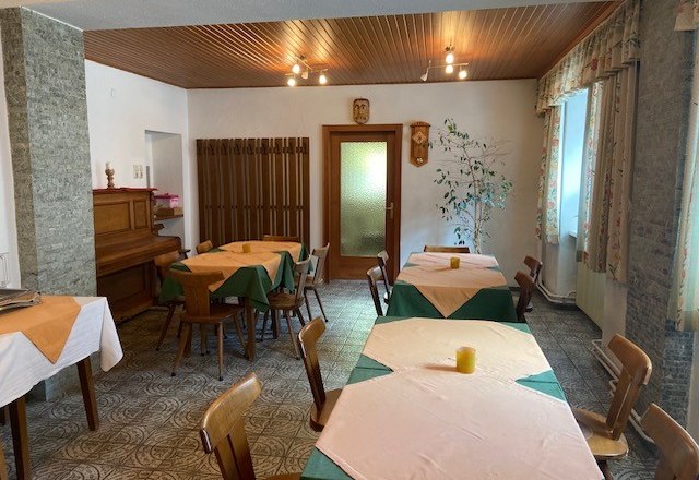 Frühstücksbereich, © Wiener Alpen Ein gemütlicher Frühstücksraum mit Holztischen, Stühlen und einem Klavier. Die Tische sind mit Tischdecken gedeckt, und es gibt Fenster mit Vorhängen.