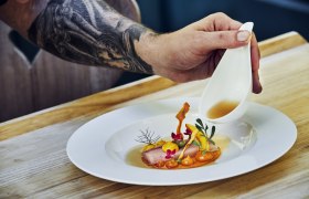 Hier isst das Auge mit, © Niederösterreich Werbung/Andreas Hofer Ein Teller mit kunstvoll angerichtetem Essen, über das eine Hand aus einer kleinen Kanne Sauce gießt.