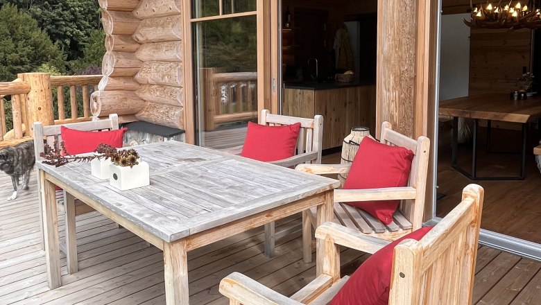 Terrasse, © RaxChalet Holzterrasse mit Tisch und Stühlen, roten Kissen und einem Hund im Hintergrund.