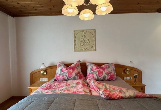 Schlafzimmer Obergeschoss, © Heidi Peer Ein gemütliches Schlafzimmer mit einem Doppelbett, floralem Bettzeug und einer Holzdecke. Eine Lampe hängt von der Decke.