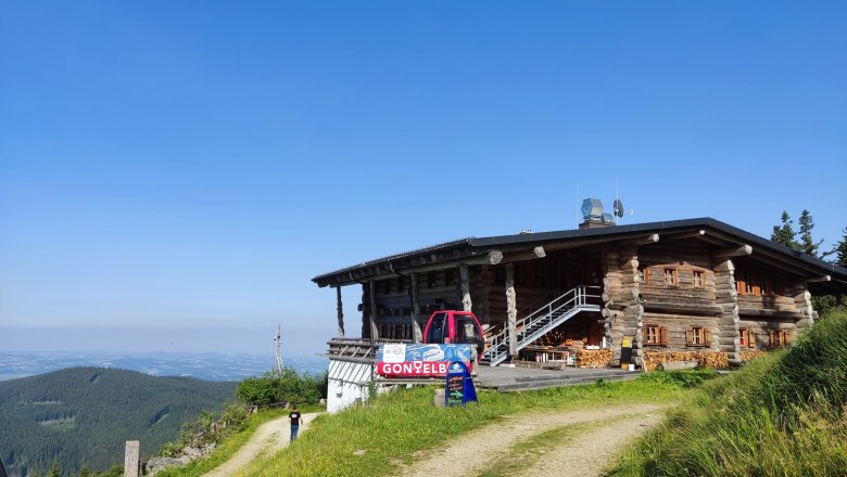 Stoa Alm, © Kulinarik zur Stoaalm/Steffen Eine Berghütte aus Holz mit einem roten Fahrzeug davor, umgeben von grünen Wiesen und einem klaren blauen Himmel.