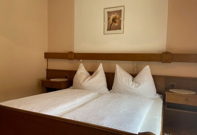 Gemütliches Doppelzimmer, © Wiener Alpen Ein gemütliches Doppelzimmer mit einem großen Bett, weißen Kissen und Decken, Holzmöbeln und einem Blumenbild an der Wand.