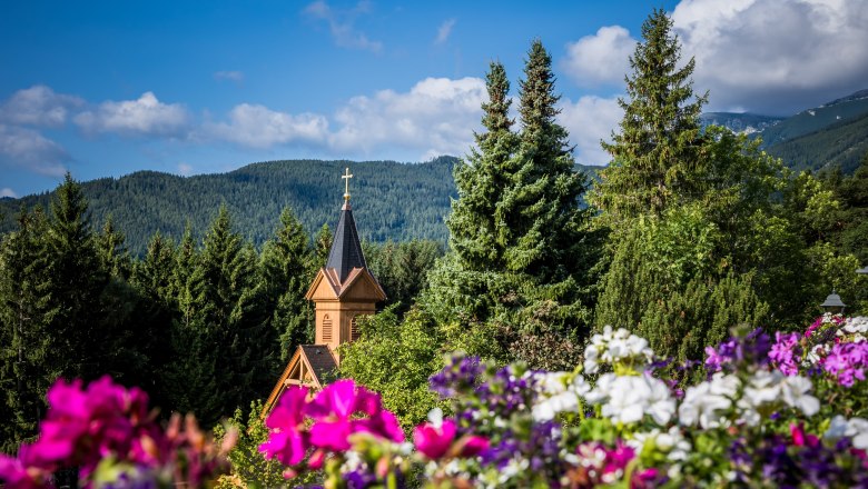 Kirche, © Knappenhof Christian Husar Kirchturm inmitten von Bäumen und Blumen vor einer Berglandschaft.