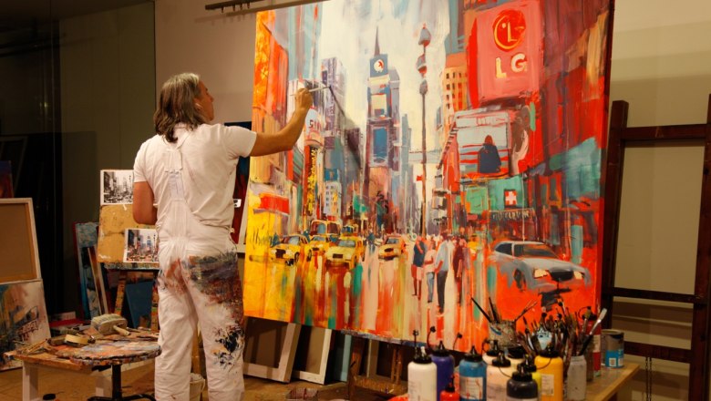 Der Künstler VOKA, © Voka Ein Künstler malt ein lebhaftes Stadtbild auf einer großen Leinwand in einem Atelier.