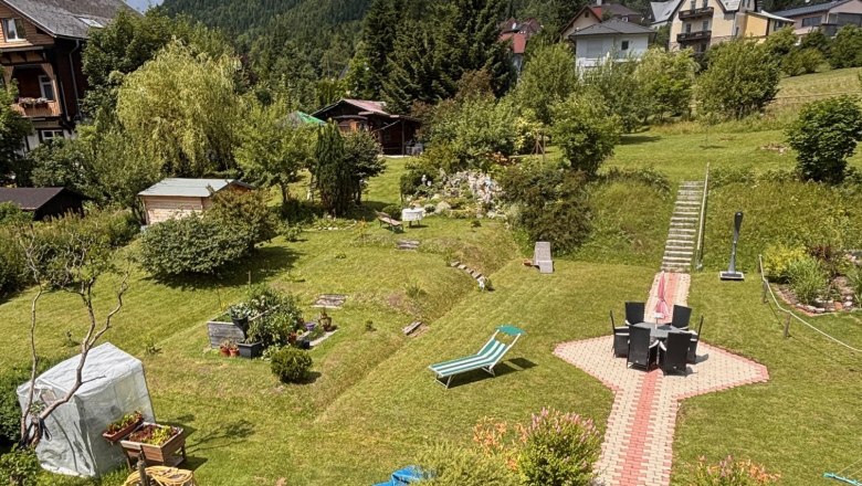 Garten, © Familie Mohr Ein gepflegter Garten mit Rasen, Gartenmöbeln und einem Weg aus Pflastersteinen. Im Hintergrund sind Häuser und bewaldete Hügel zu sehen.