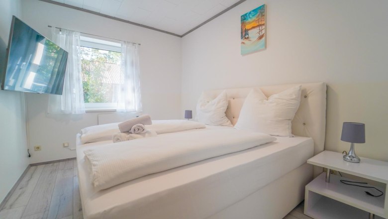 Schlafzimmer, © Michaela Narath Modernes Schlafzimmer mit Doppelbett, weißer Bettwäsche, Wandbild, Fenster mit Vorhängen und Flachbildfernseher.