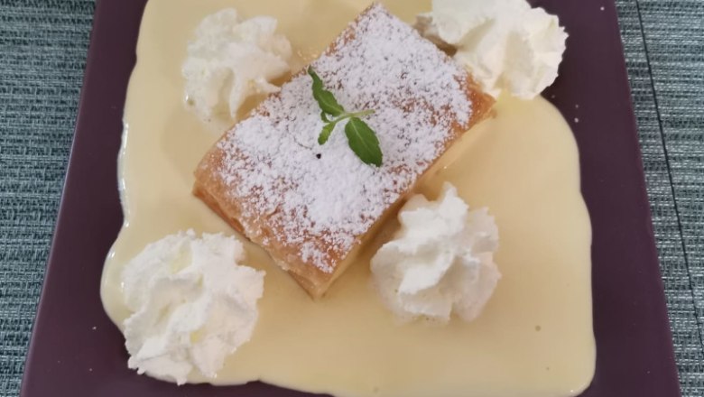 Cafe Elli, © Cafe Elli Ein Dessert auf einem lila Teller: Ein Stück Topfenstrudel mit Puderzucker, umgeben von Vanillesoße und Sahnehäubchen, garniert mit einem Minzblatt.