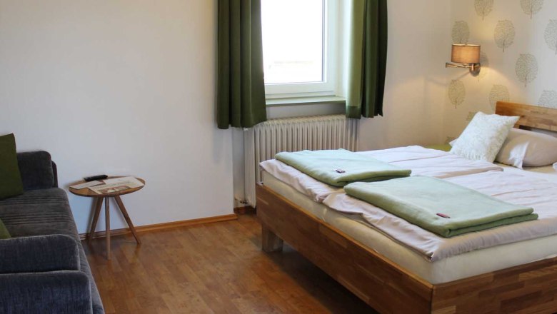 Doppelzimmer, © Magdalena Donhauser Schlafzimmer mit Doppelbett, Fenster mit grünen Vorhängen, Sofa, Tisch, Tapete und einer Wandlampe.