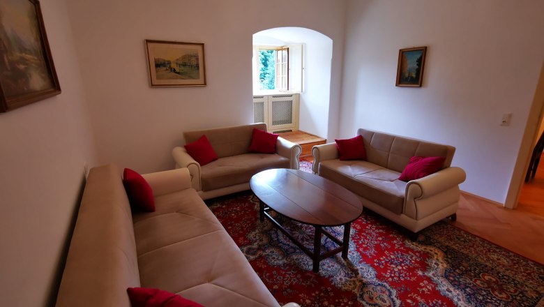 Wohnzimmer im Appartement Josef, © Herrnhof Wohnzimmer mit drei Sofas, rotem Teppich und Gemälden an den Wänden.