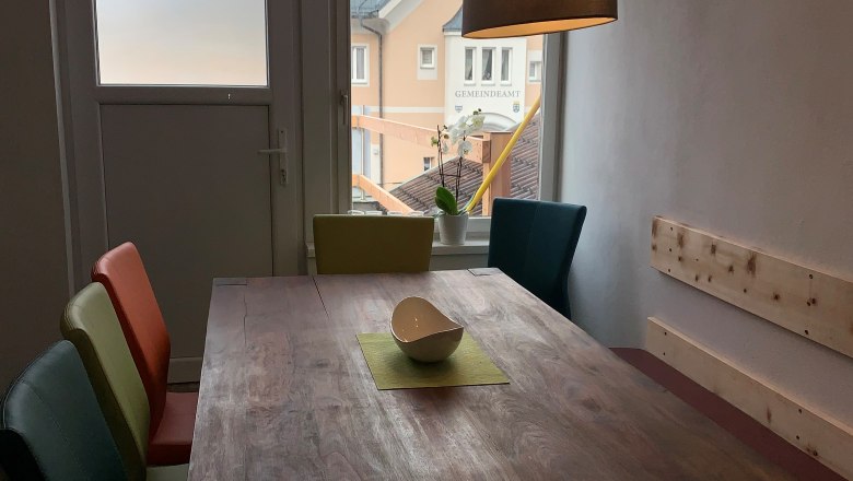 Küche-Essbereich, © Seidl Essbereich mit Holztisch, bunten Stühlen und Hängelampe, Fenster mit Blick auf Gebäude.