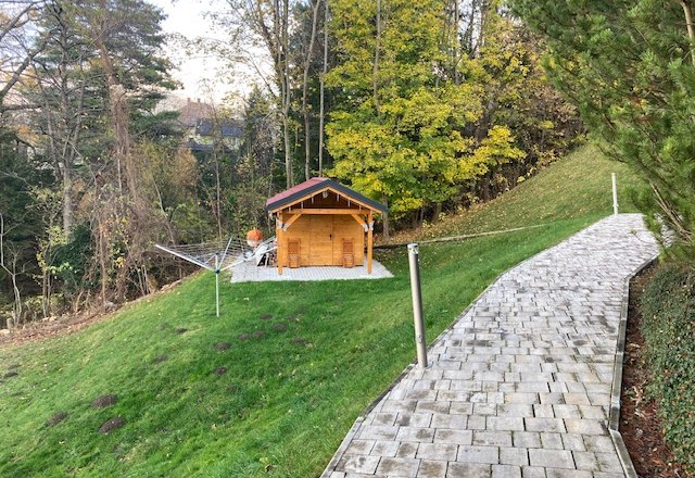 Gartenbereich, © Thomas Ruzicka Ein Garten mit einem kleinen Holzhaus, umgeben von Bäumen und einem gepflasterten Weg.