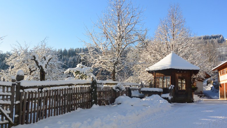 Winterstimmung, © Erika Kronaus Verschneite Landschaft mit Holzhütte, Holzzaun und Bäumen unter blauem Himmel.