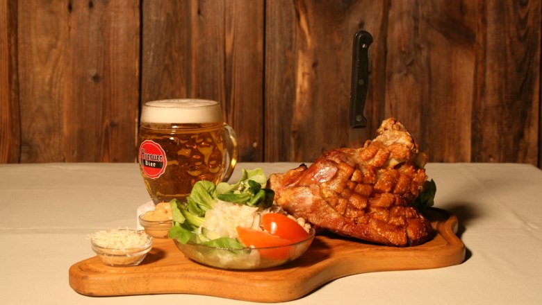 Stelzen Familie Kamper, © Familie Kamper Ein Holzbrett mit Schweinshaxe, Salat und Bierkrug vor Holzwand.