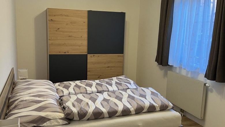 Doppelzimmer, © Andrea Tauchner Ein gemütliches Doppelzimmer mit einem Holzbett, einem Kleiderschrank und einem Fenster mit Vorhängen.