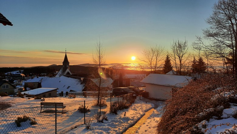 Sonnenaufgang von der Belle Air Logde im Winter, © Christoph Gierlinger Winterlicher Sonnenaufgang über einer verschneiten Landschaft mit Kirche und Häusern.