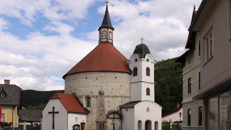 Pfarrkirche Scheiblingkirchen, © Steindy, CC BY-SA 3.0 Pfarrkirche Scheiblingkirchen mit rundem Turm und rotem Dach, umgeben von Gebäuden und bewölktem Himmel.