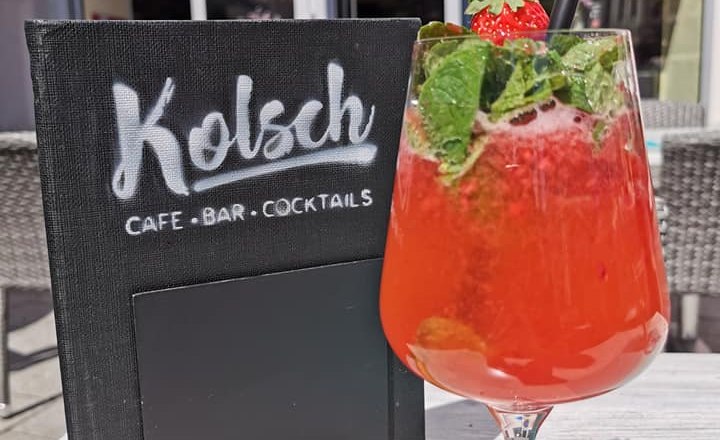 Café und Cocktail Bar, © Facebook Das Kolsch Ein roter Cocktail mit Minze und Erdbeere vor einem Café mit Schild 'Kölsch Café Bar Cocktails'.