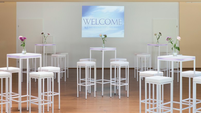 Seminar at the Parkhotel Hirschwang, © missionINGE.com Seminar room with white bar tables and stools, vases of flowers and a screen with the inscription 'Welcome'.