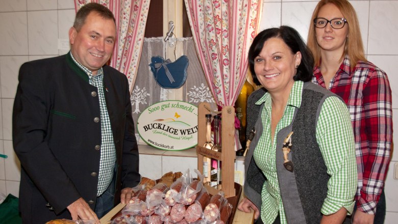 Hofladen Karnthaler, © Hans Tomsich Drei Personen stehen in einem Hofladen vor einem Tisch mit Wurstwaren. Im Hintergrund ein Schild mit der Aufschrift 'Bucklige Welt'.