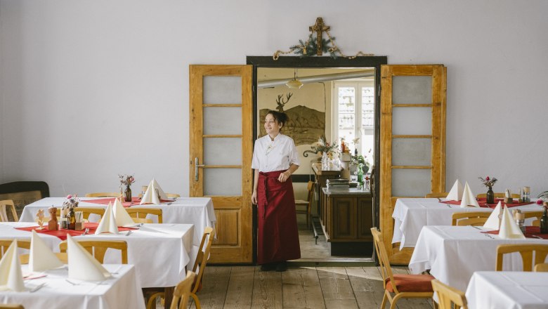 Kräuterwirtin Gerda Stocker Saal zum Genießen und Feiern, © Wiener Alpen/Sophie Menegaldo Eine Frau in einer weißen Bluse und roter Schürze steht in einem festlich gedeckten Raum mit Holzboden und weißen Tischdecken.