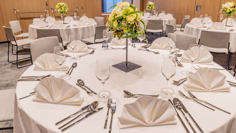 Gedeckte Tische, © Hilton Garden Inn Wiener Neustadt Rund gedeckter Tisch mit weißer Tischdecke, Besteck, Gläsern und Blumenarrangement in einem eleganten Raum.