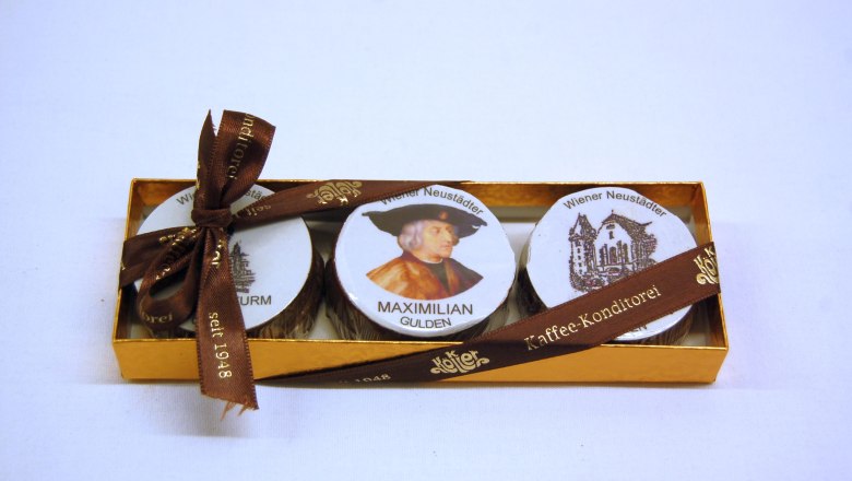Der Wr. Neustädter Maximilian-Gulden, © Konditorei Köller Eine goldene Schachtel mit drei Pralinen, verziert mit einem Bild von Maximilian und Wiener Neustadt Motiven, gebunden mit einem braunen Band.