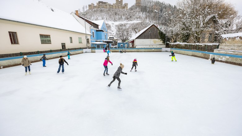 Eislaufplatz Kirchschlag, © Wiener Alpen, Martin Fülöp Menschen eislaufend auf einem Platz in Kirchschlag, im Hintergrund auf einem Hügel eine Burgruine.