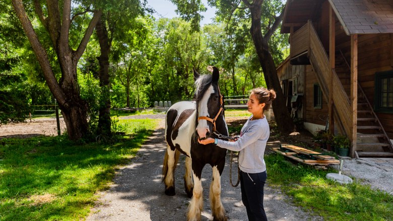 Pferdewelt Reichenau, © Wiener Alpen / Christian Kremsl Eine Frau steht neben einem schwarz-weißen Pferd auf einem Hof mit Bäumen und einem Holzgebäude im Hintergrund.