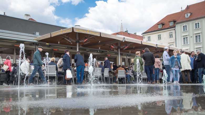 der Springbrunnen, © WNSKS Menschen sitzen und stehen um einen Springbrunnen auf einem Platz mit Café.