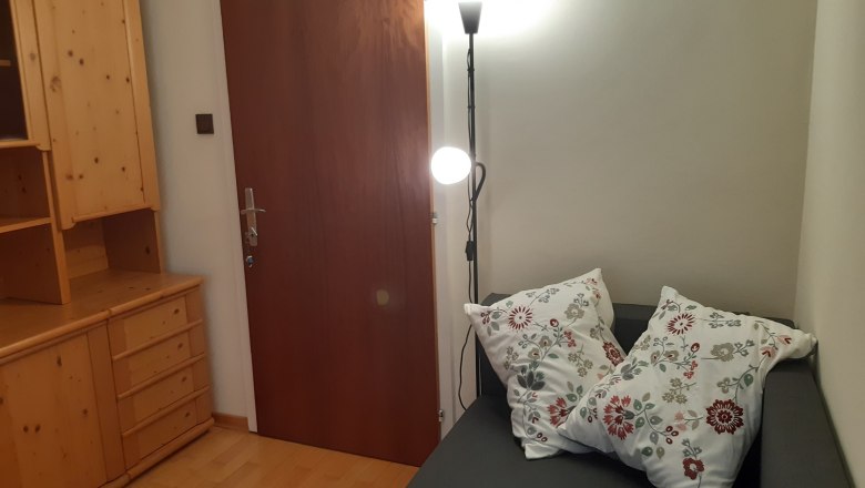 Abendstimmung - Zimmer 3, © Kainz-Waldinger Ein gemütliches Zimmer mit einer grauen Couch, dekoriert mit zwei bestickten Kissen, einem Holzschrank und einer Stehlampe neben einer Holztür.