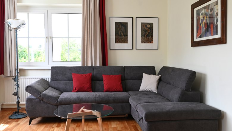 Wohnzimmer, © Christine Kuppelwieser Modernes Wohnzimmer mit grauem Sofa, roten Kissen, Glastisch und Kunstwerken an der Wand.