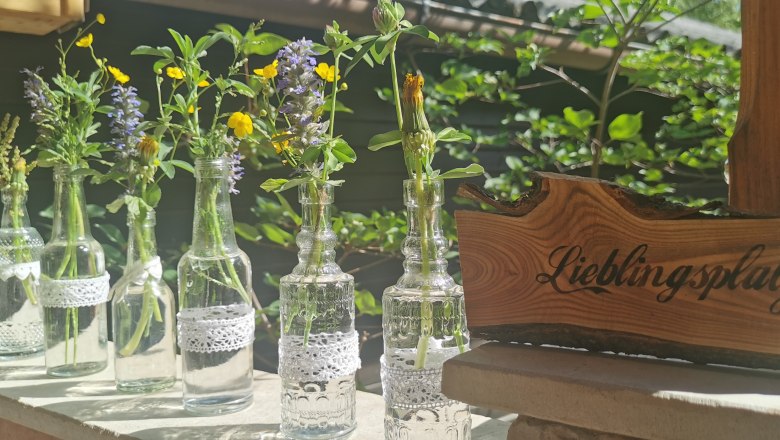 Dekoration in Mei Kuchl, © Anita Brem-Hofer Kleine Glasflaschen mit Wiesenblumen und ein Holzschild mit der Aufschrift 'Lieblingsplatz'.
