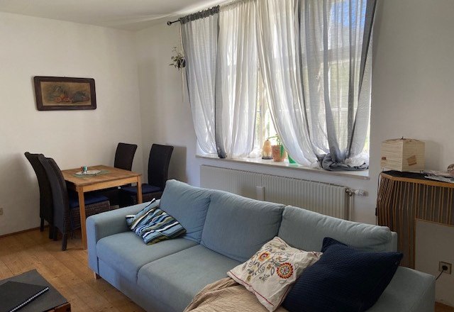 Wohnzimmer, © Wiener Alpen Ein Wohnzimmer mit einem blauen Sofa, einem Holztisch mit Stühlen und einem Fenster mit weißen Vorhängen.