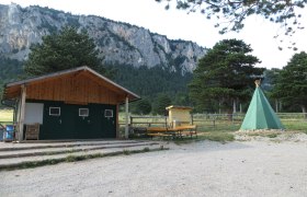 Zeltplatz Hohe Wand Blick Camp, © Naturpark Hohe Wand Ein Zeltplatz mit einem grünen Tipi, einer Holzhütte und Bänken vor einer Felswand und Bäumen.