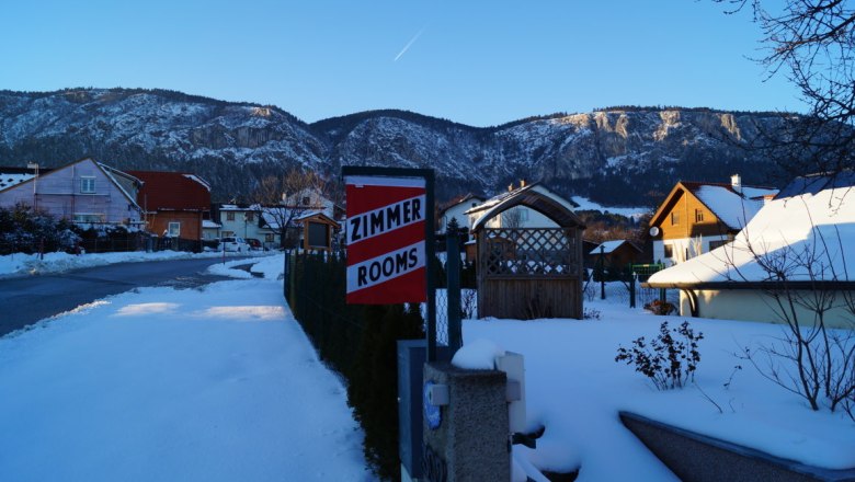 Aussicht vor dem Haus, © Reep Winterliche Landschaft mit schneebedeckten Häusern und Bergen im Hintergrund. Ein Schild mit der Aufschrift 'Zimmer Rooms' ist sichtbar.