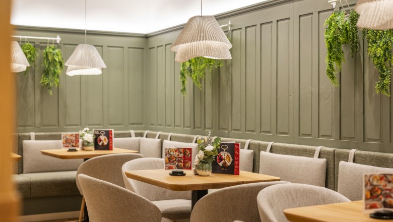 Cafe area - Bakery Koll, © Michaela Begsteiger, Koll GmbH Cozy café area with wooden tables, grey chairs and green wall panelling. Hanging plants and lamps create an inviting atmosphere.