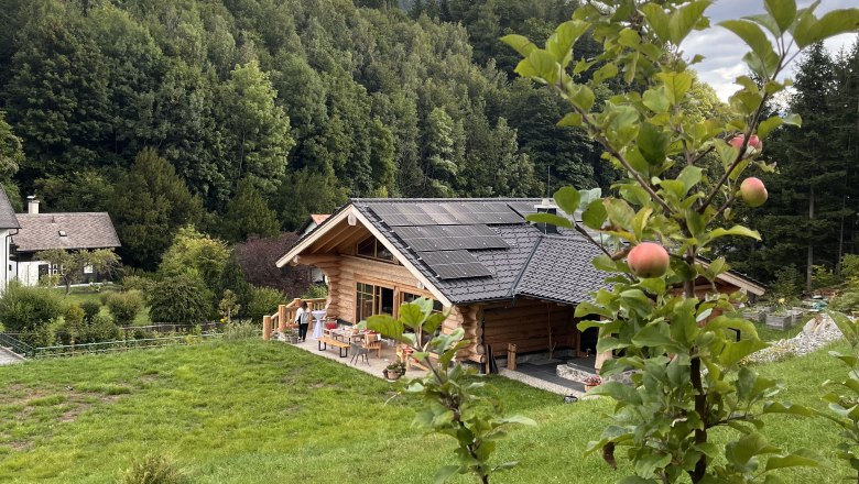 Hausansicht RaxChalet, © RaxChalet Ein Holzchalet mit Solarpanelen auf dem Dach, umgeben von grüner Natur und einem Apfelbaum im Vordergrund.